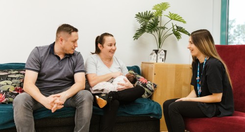 Image of a mother breast feeding her baby while sat with the father and a healthcare professional.